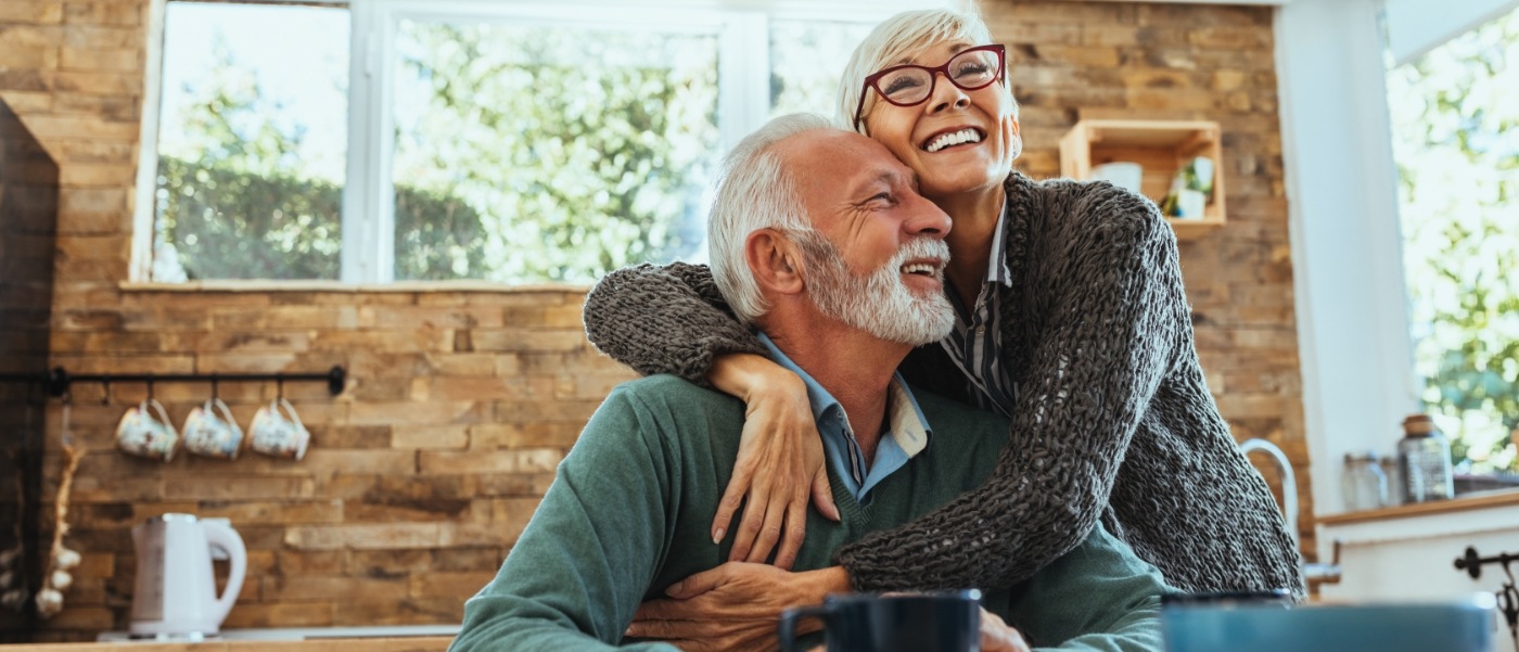 Senior man and woman hugging at kitchen table and smiling with dental implants in Califon